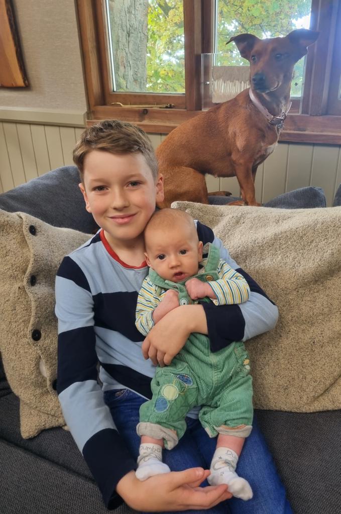 Zachery smiling whilst holding a baby, with a curious dog in the background